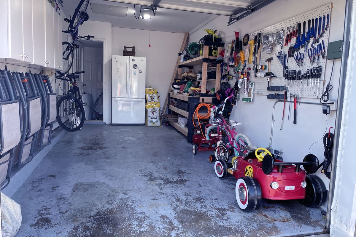 Interior of a narrow garage with lots of stored tools, bike parts, and a refrigerator on the back