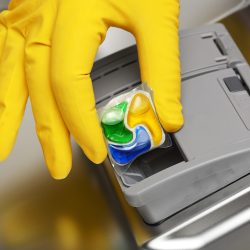 Close-up of hand in yellow rubber glove putting detergent tablet into dishwater - Dishwasher Pods Vs. Liquid - Which To Choose For Your Kitchen