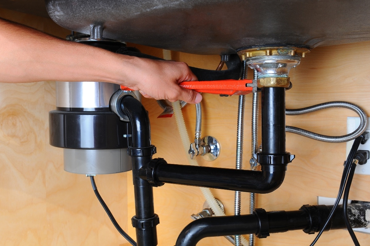 Closeup of a plumber using a wrench to tighten a fitting beneath a kitchen sink -Can I Replace Garbage Disposal Myself