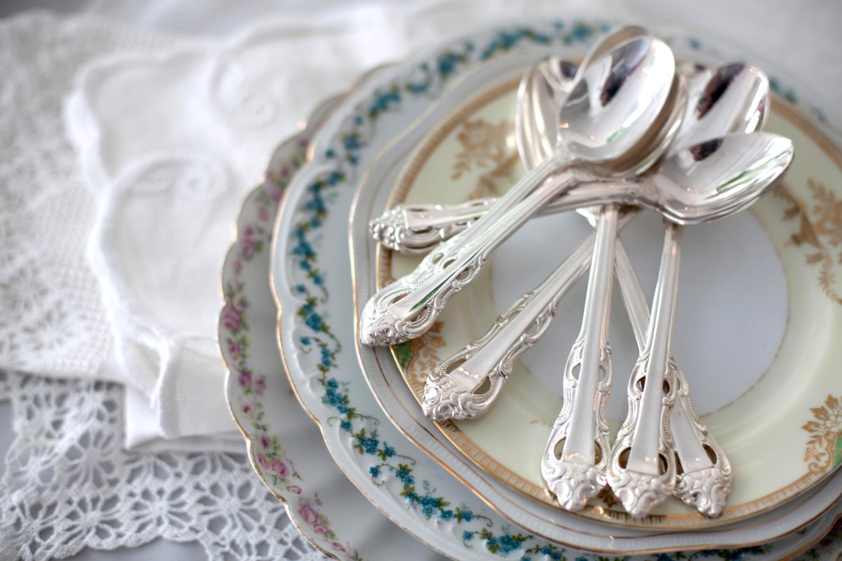 A stack of vintage floral plates with silver teaspoons on a table near lace doilies