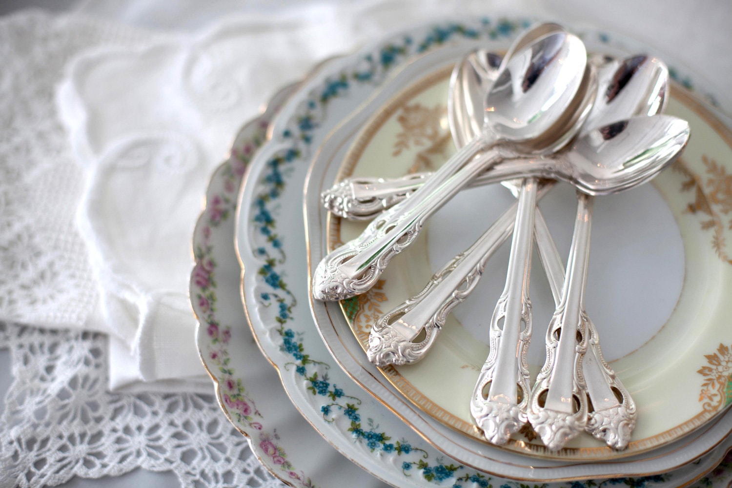 A stack of vintage floral plates with silver teaspoons on a table near lace doilies.