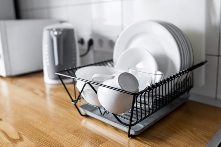 Budget and lightweight antimicrobial dish drainer with drain board at modern scandinavian kitchen. Dish rack holds many dishes and cups against wooden countertop, white wall tiles, sink and faucet, What's The Best Dinnerware For Both Microwave & Dishwasher?