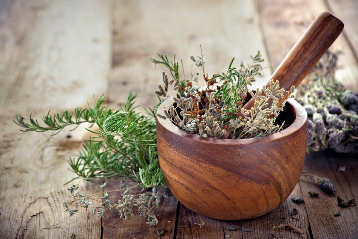 Fresh herbs inside a wooden mortar and pestle bowl on a wooden plank table with knotholes and nails. The herbs have multiple colors and textures. The bowl has a swirl pattern and is surrounded by piles of additional fresh herbs. - How To Season A Wooden Mortar And Pestle [Inc. Maintenance Care]