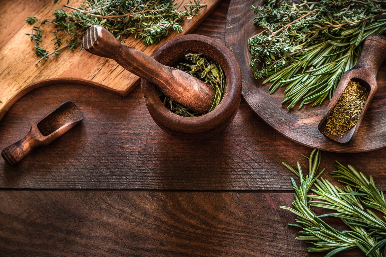 High angle view of natural aromatic herbs like dill, thyme, oregano and rosemary on wooden mortar and over dark brown wooden table. Predominant colors are brown and green. 