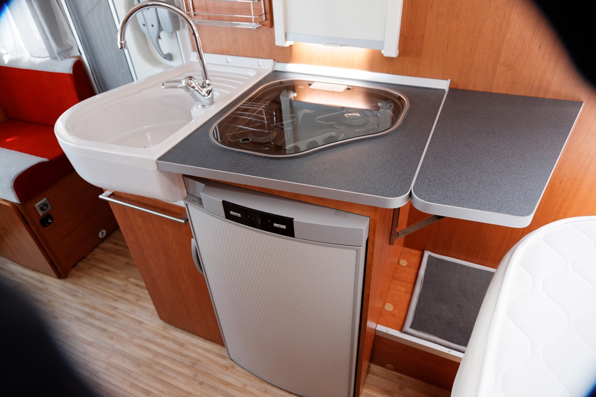 Interior of motor home, camping, motorhome, ARV, showing a side kitchen with glass covered hob, sink. Fridge under cabinet.