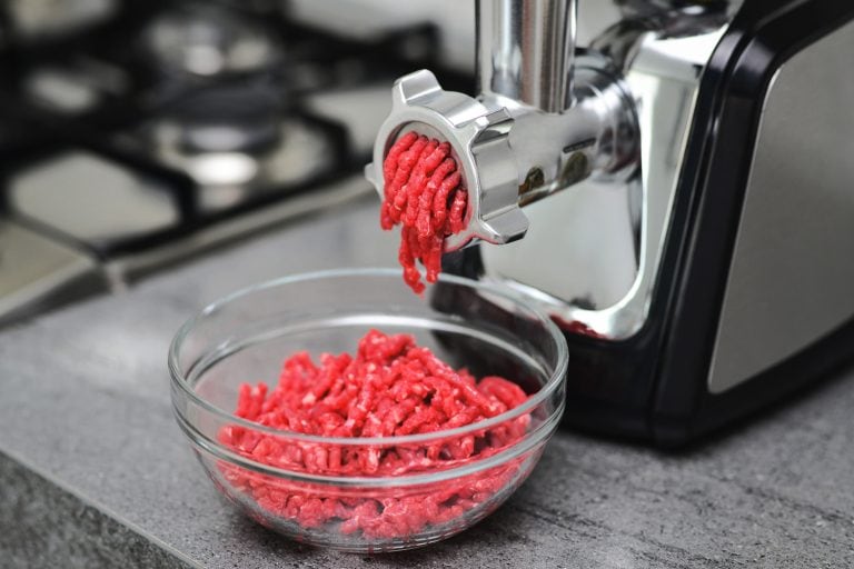 Meat grinder with fresh forcemeat on kitchen table. Side view of a meat grinder and minced meat falling into a glass bowl. Selective focus. - Can You Grind Cooked Meat In A Meat Grinder Or Blender
