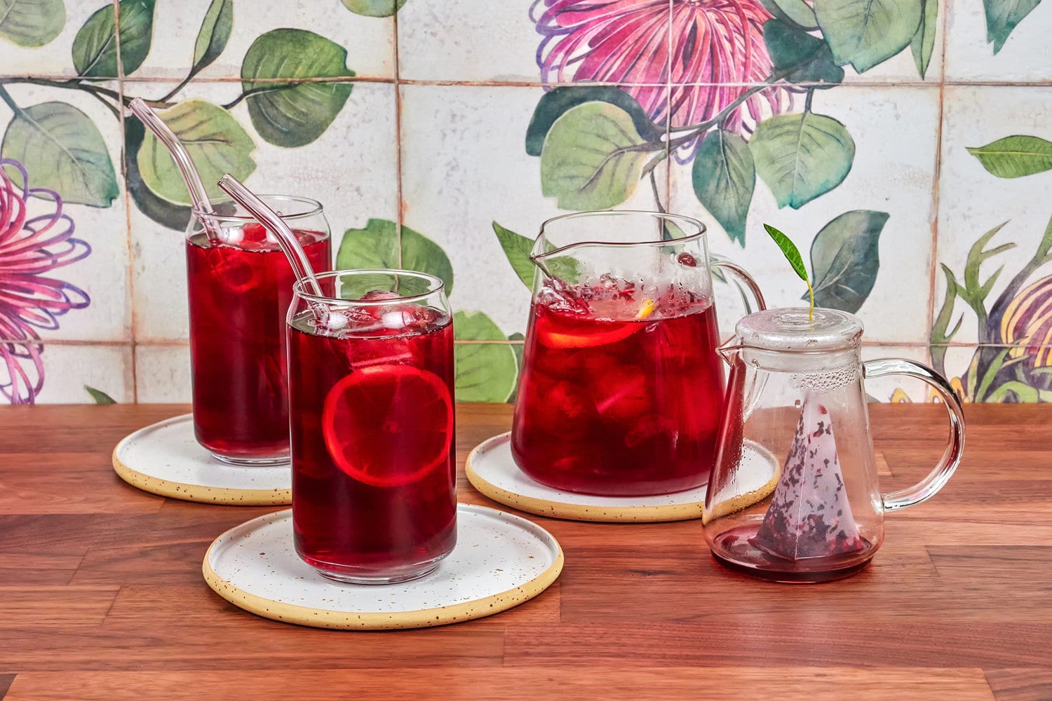 Raspberry ice tea in a special glass teapot, with two ice tea glasses and borosilicate straws, on a walnut table and ceramic tiles in the background.