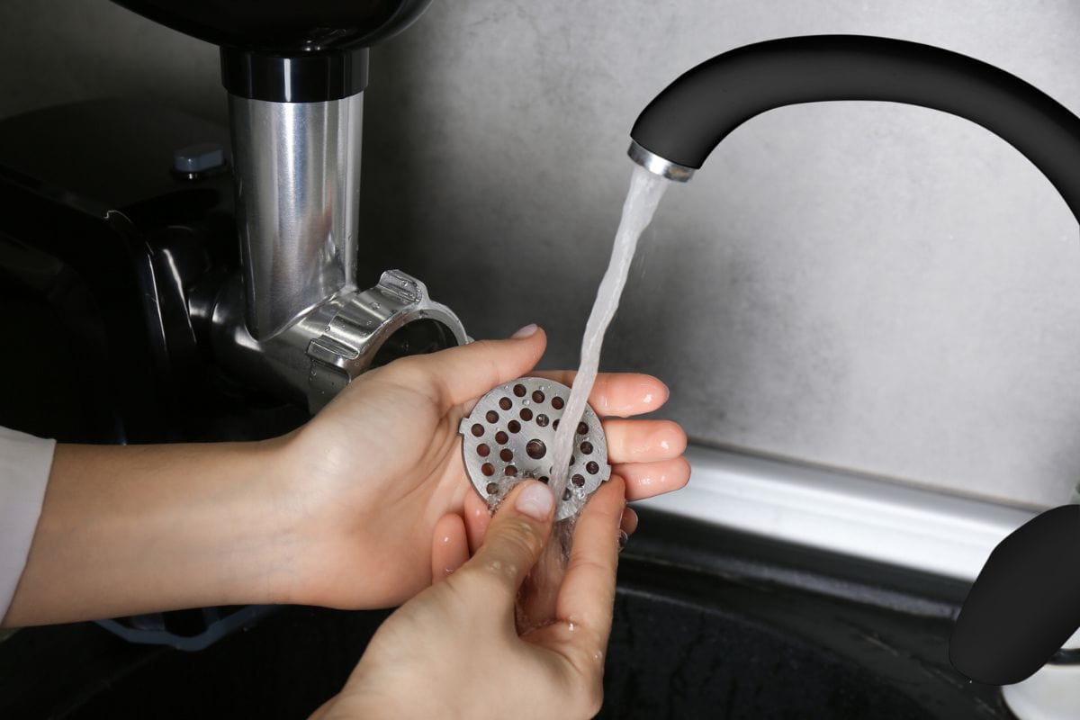 Woman washing cutting plate of electric meat grinder under tap water in kitchen sink indoors, closeup