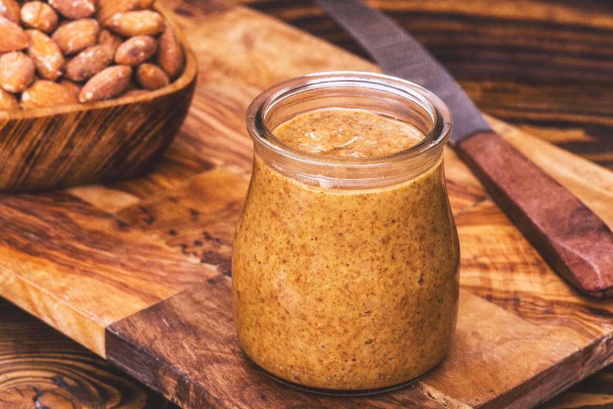 A jar of homemade raw organic almond butter with a wood bowl of fresh salty almonds and knife on the side on the wood board
