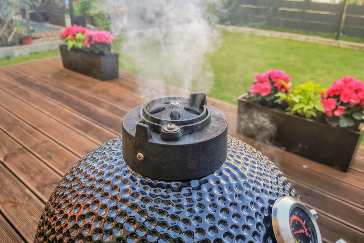 A kamado egg type barbeque grill standing on a living house terrace with a smoke coming from it's chimney, used as a cold smoke house