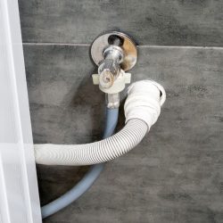 Dirty drain and water hoses for the washing machine running into a wall covered with gray concrete tile. Design options in a comfortable modern bathroom. - Where To Drill Hole For Dishwasher Drain Hose?
