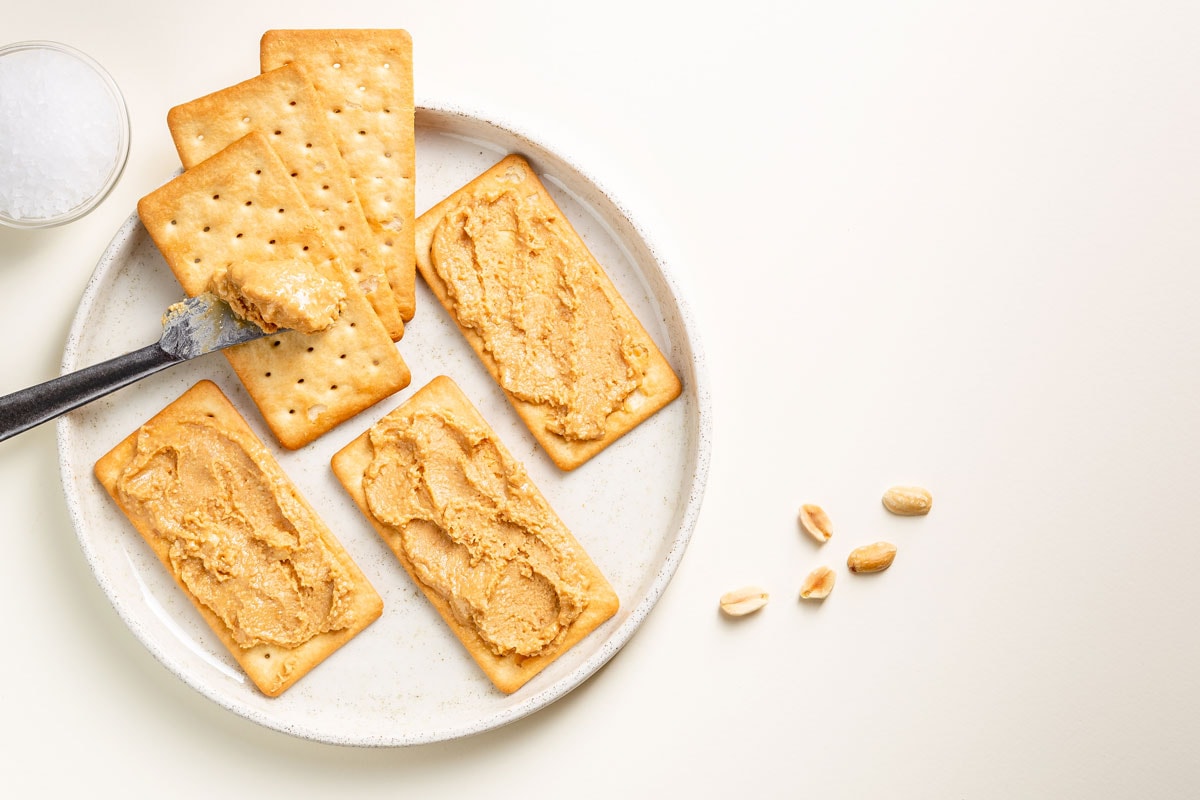 Peanut butter spread on soda crackers on the plate seen from above with knife and salt