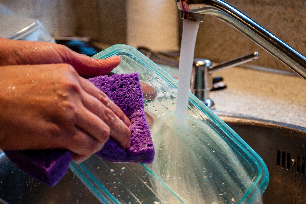 Woman washing a plastic food storage container with a sponge in kitchen sink
