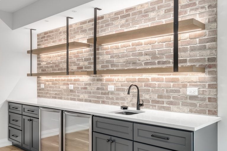 A basement wet bar with a white granite countertop, grey cabinet, lighting under the shelves mounted to exposed brick, and built-in refrigerators - What Size Sink Do I Need For A Basement Bar