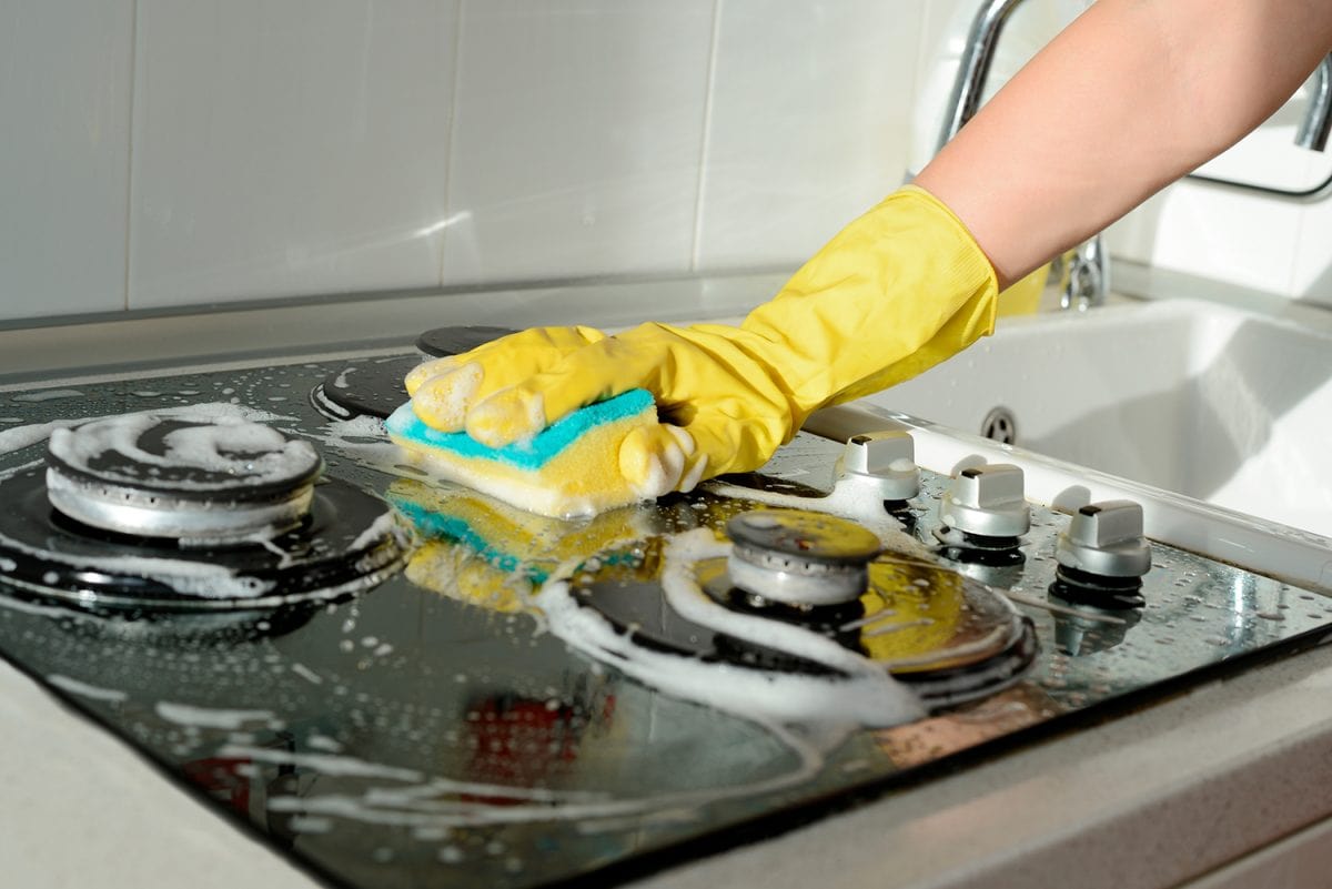 A hand in a yellow rubber glove washes a gas stove on a sunny day