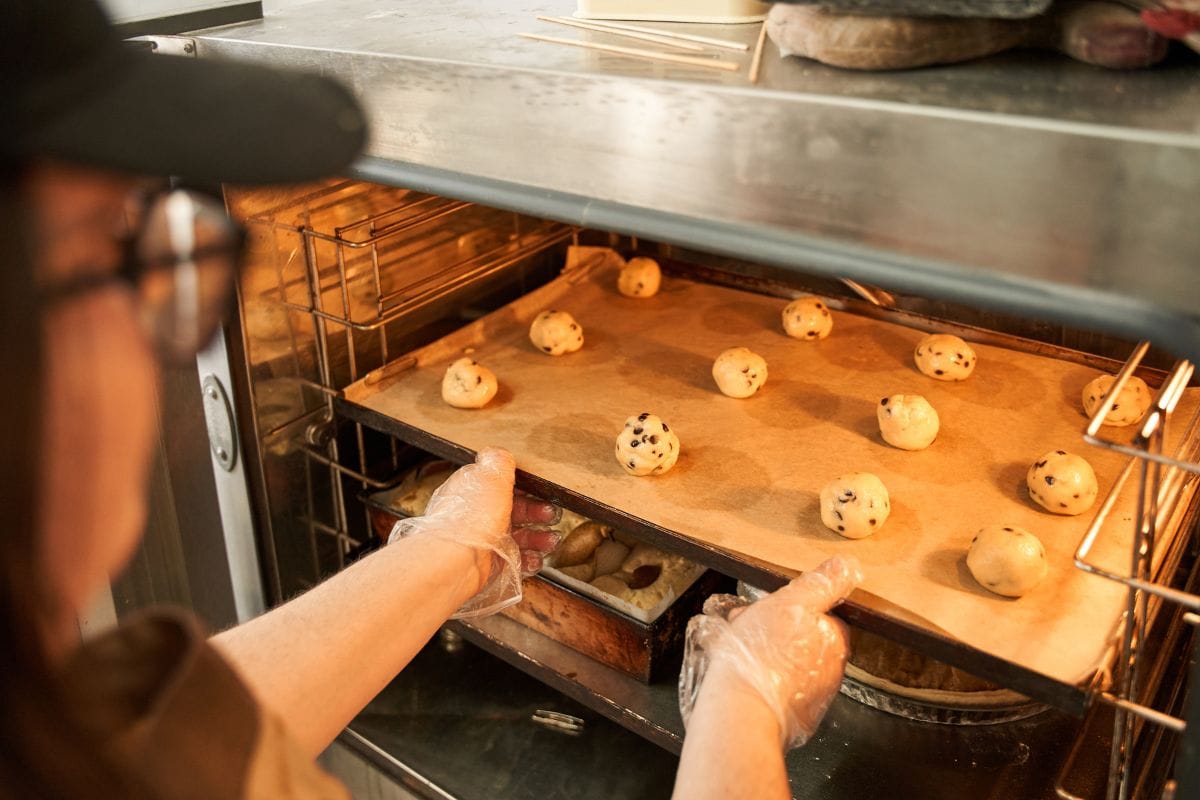 Baker putting tray with balls from the dough to the oven while making cookies 