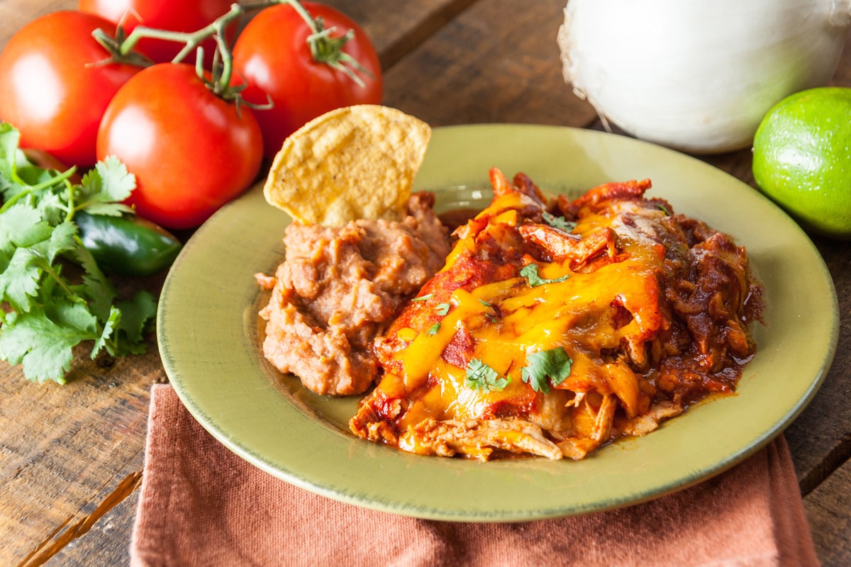 Chicken Enchilada Mexican Food Casserole Served On Rustic Green Plate On Vintage Wood Table Background, With Tomato, Jalapeno, Onion And Lime Fresh Ingredients.