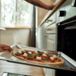 Cropped shot of man, professional cook making pizza at home. Man in apron putting raw pizza in modern oven for baking. Hobby, lifestyle. Selective focus. Side view. Web Banner, How Close Can A Pizza Oven Be To The House?