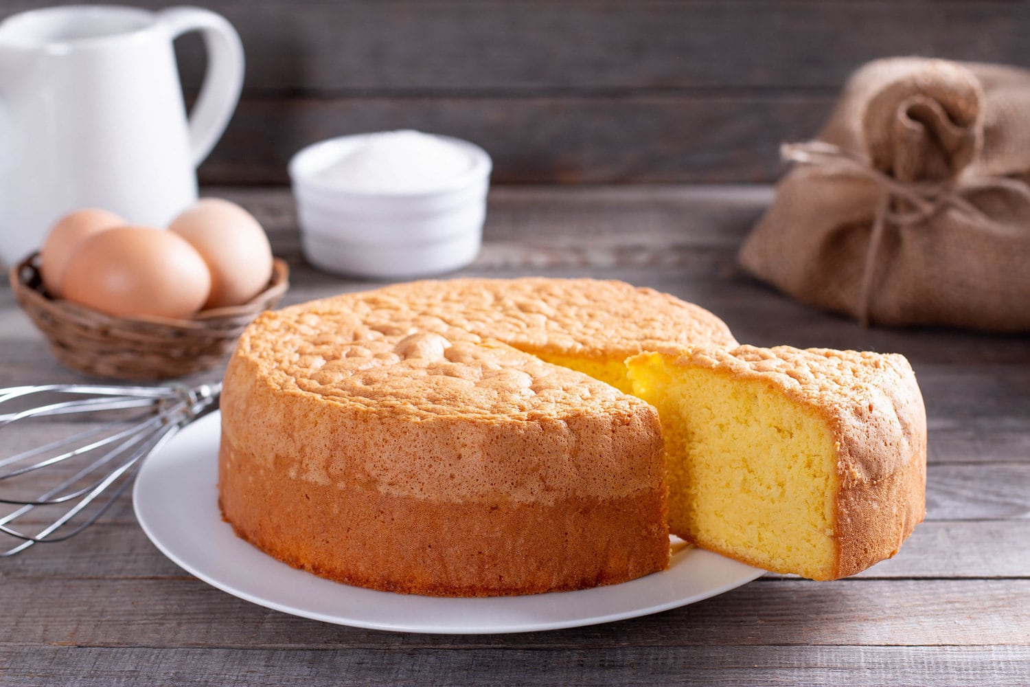 Homemade round sponge cake or chiffon cake on white plate so soft and delicious with ingredients eggs, flour, milk on wood table. Homemade bakery concept for background and wallpaper.