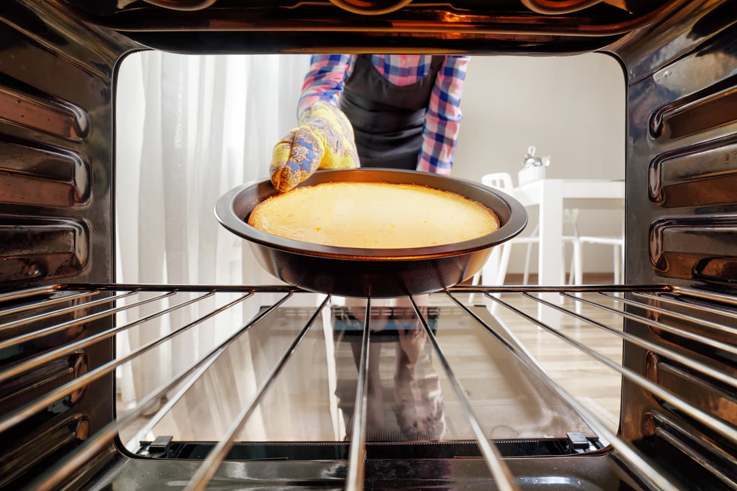 Housewife taking cheesecake out of oven in kitchen. View from inside of the oven. Woman wearing black apron and colorful oven mitt. 