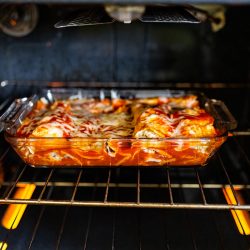 Inside oven with rack and homemade Mexican food enchiladas in baking tray glass dish cooking with tomato sauce, tortilla and cheese, Should You Bake Enchiladas Covered Or Uncovered?