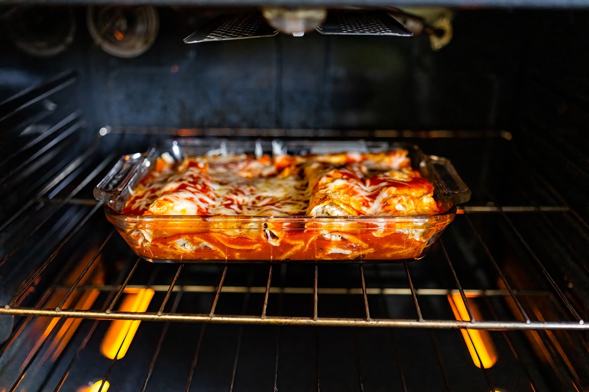 Inside oven with rack and homemade Mexican food enchiladas in baking tray glass dish cooking with tomato sauce, tortilla and cheese, Should You Bake Enchiladas Covered Or Uncovered?