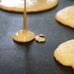 Pancake batter baking mix being poured from a bowl onto a hot electric griddle, How Thick Should Pancake Batter Be?
