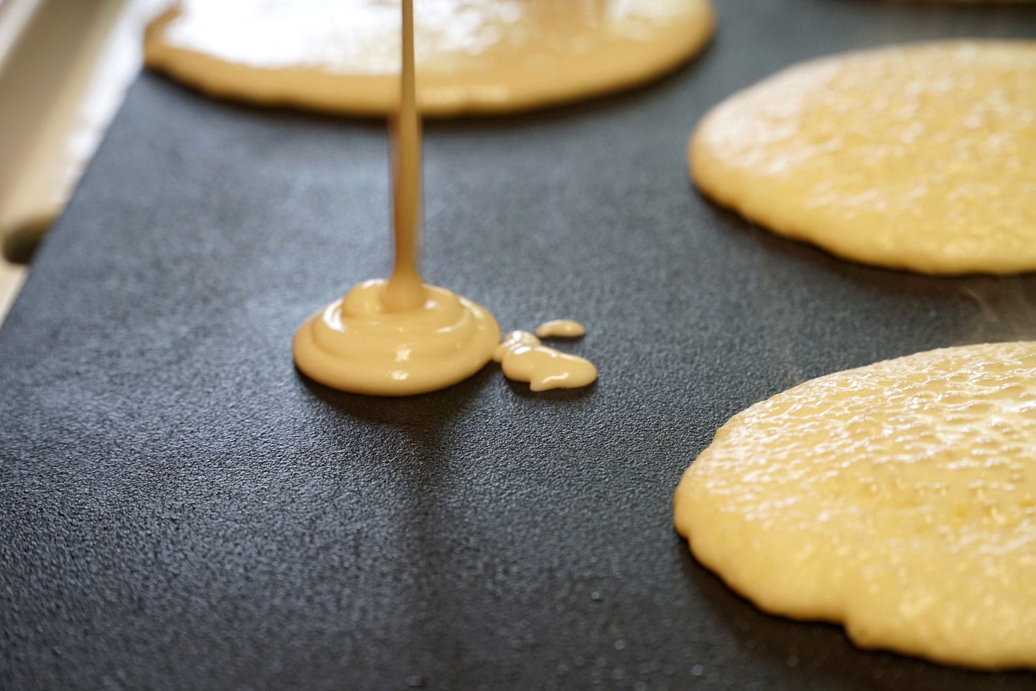 Pancake batter baking mix being poured from a bowl onto a hot electric griddle