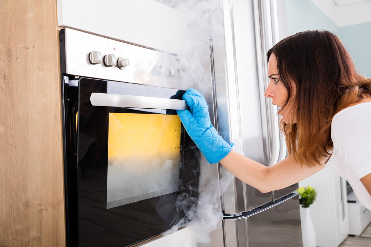 Shocked Young Woman Looking At Smoke Coming Out Of Oven In Kitchen