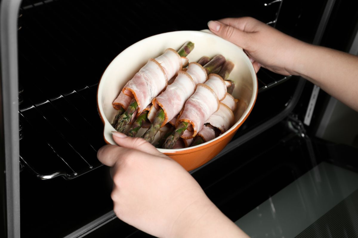 Woman putting ceramic baking dish with bacon wrapped asparagus in oven, closeup