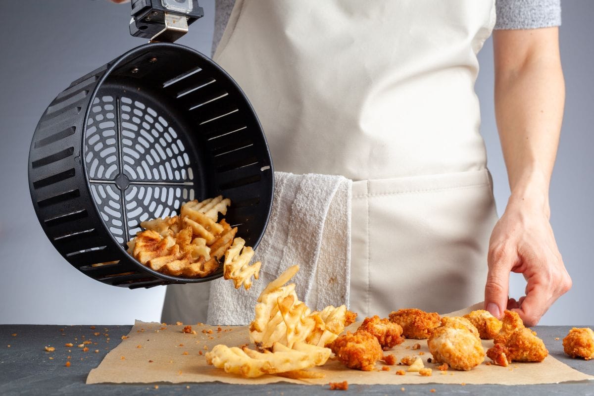 A woman is dumping fresh made potato waffle fries from basket onto a countertop together with chicken nuggets. She fried them in air fryer using very little fat. A healthy homemade convenient snack.
