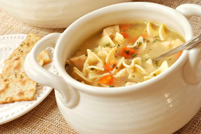 Chicken noodle soup in cream colored ceramic bowl with handles. Plate of crackers and soup tureen in background. Closeup with shallow dof., 11 Soups for Winter that Will Warm Your Heart