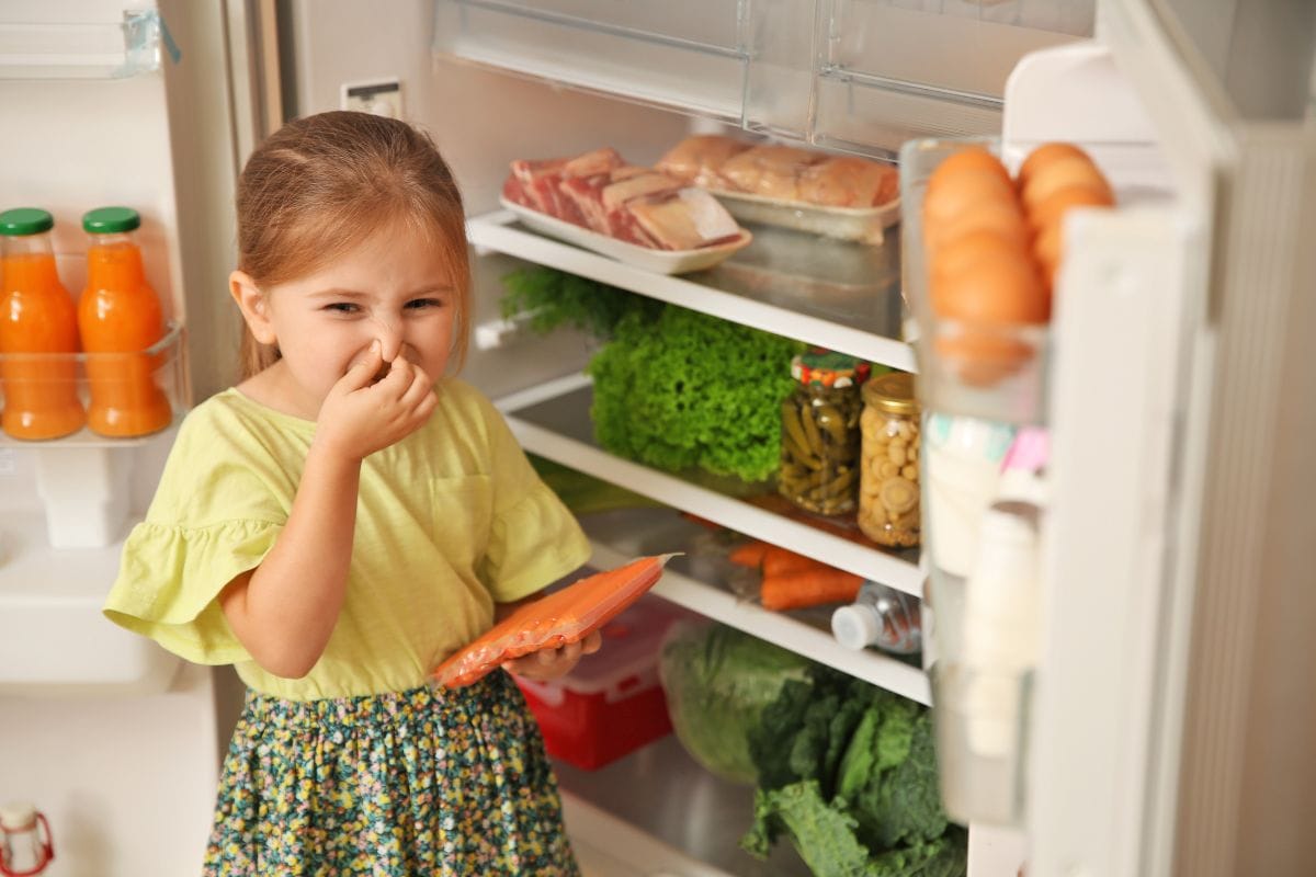Cute little girl with bad smelling sausages near refrigerator at home
