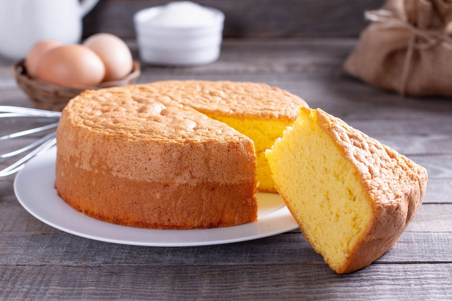 Homemade round sponge cake or chiffon cake on white plate so soft and delicious with ingredients eggs, flour, milk on wood table. Homemade bakery concept for background and wallpaper.