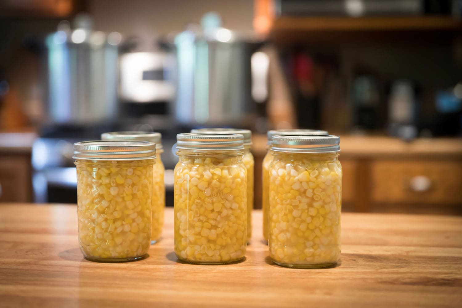 Pint jars of corn sitting on the counter ready to go in pressure canners that are sitting on the stove in the background.
