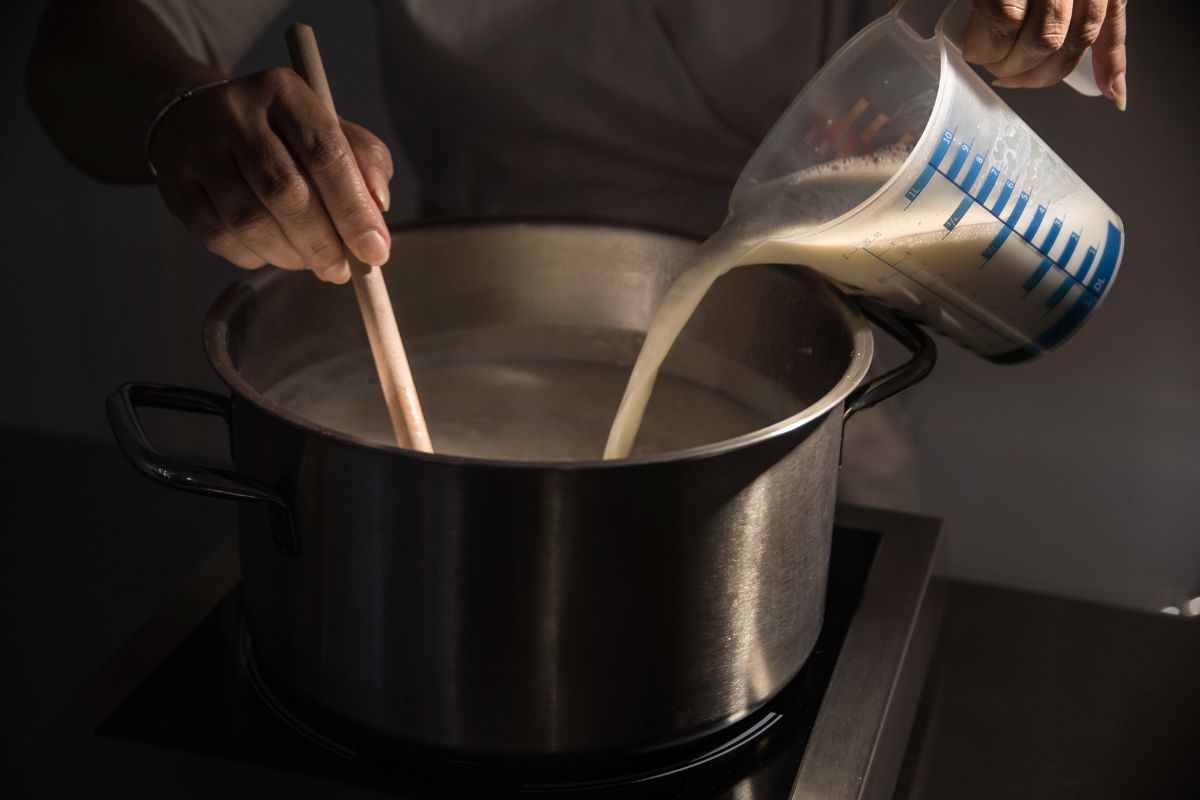 Pouring milk from beaker into metal pot and mixing it with rice to cook rice pudding