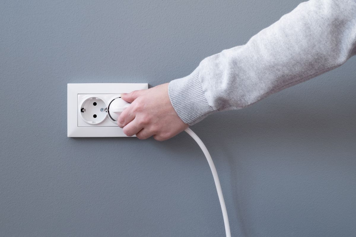 Hand plugging in an electric cord into a white plastic socket or european wall outlet on grey plaster wall. Closeup of a woman's hand inserting an electrical plug into a wall socket.