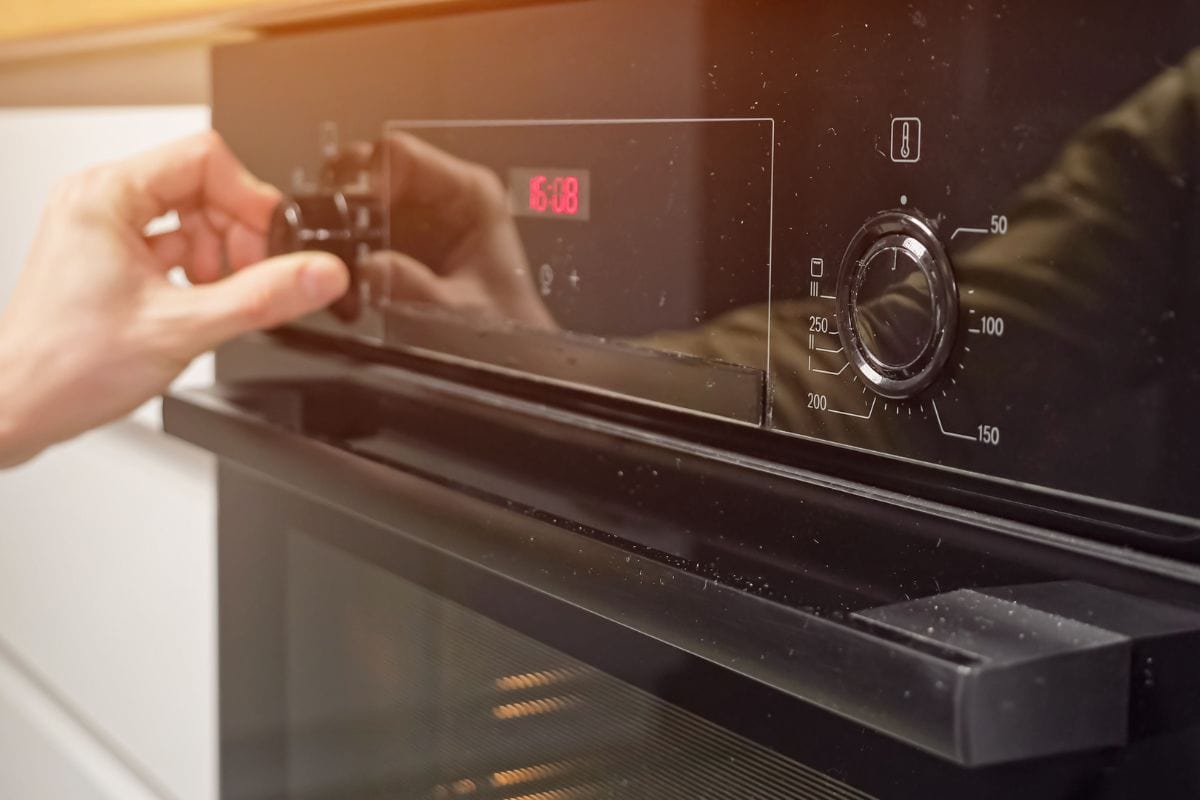 Man adjusts temperature and sets operating mode on modern oven in kitchen. Housekeeper prepares dinner using cooking appliance at home, sunlight.