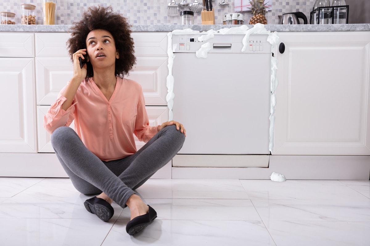 Sad Woman Calling Technician On Cellphone To Fix Dishwasher With Foam Coming Out From It
